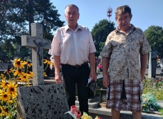 Dwóch Panów na cmentarzu przy jednym z grobów, na grobie znajdują się wieńce z kwiatów i znicz